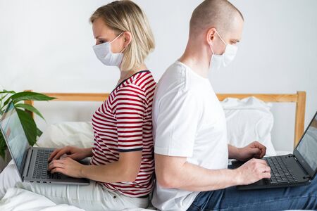 family working remotely from home siting back to back on bed. Quarantined couple coronavirus in medical masks .Stay home safe. Distance learning, education and work. Order food products online.の写真素材
