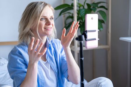 Woman blogger talking with followers, live streaming, looking to smartphone screen sitting on bed. Making video call to friends and parents.の写真素材