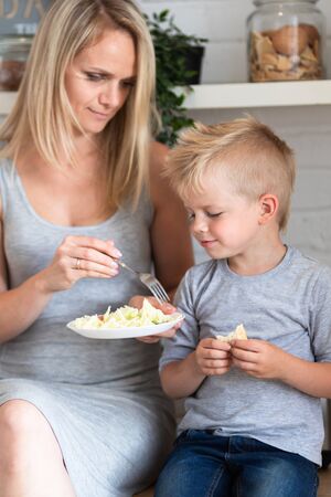 blonde family mother and son eating healthy food in kitchen at home, green salad on plates.の写真素材