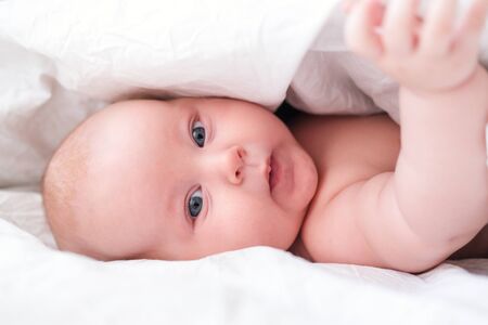 Adorable smiling baby boy under white blanket in bedroom. Newborn child relaxing in bed. Family...