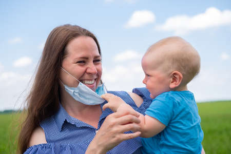 Happy mother in protective medical mask holds her little son on arms in nature at summer. End of coronavirus, childhood and family conceptの写真素材
