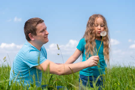 father and daughters spending time. Dad and daughters playing together on field green grass at summer. girl blowing on a dandelionの写真素材