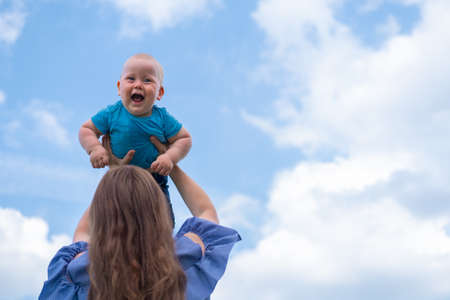 Mom lifting baby high into blue cloudy sky. Boy smiling and looking at camera. Happy childhood and family concept. Place for text.の写真素材