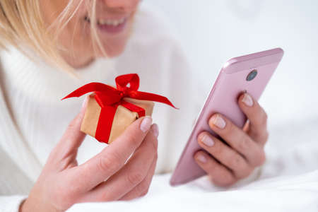Christmas online shopping. Female buyer makes order on mobile phone. Woman buy presents, prepare to xmas, gift box in hand. Winter holidays sales.の写真素材