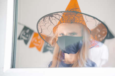 Portrait sad blonde girl in witch costume wearing face masks looking through window preparing for...
