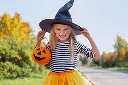 Halloween kids. Portrait smiling girl in witch hat with pumpkin candy bucket. Funny kids in carnival costumes outdoors.の写真素材