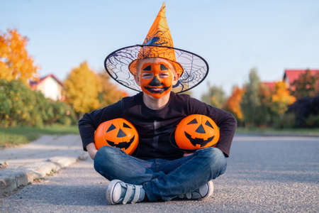 Halloween kids. Laughing boy with pumpkin face mask in witch hat with two candy buckets sitting on street.の写真素材