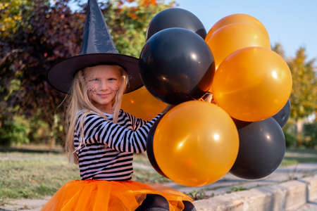 Halloween kids. Portrait smiling girl in witch hat with orange and black balloons. Funny kids in carnival costumes outdoors.の写真素材