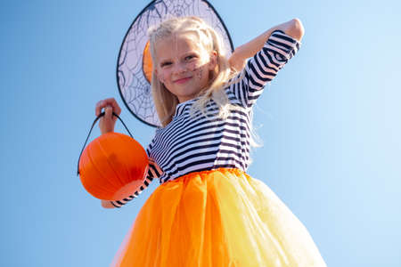 Halloween kids. Portrait Laughing girl in witch costume hat with pumpkin candy bucket on street.の写真素材