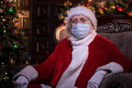 Santa Claus in medical mask. Portrait positive Santa sitting near Christmas tree. Happy New Year Covid 2021.の写真素材