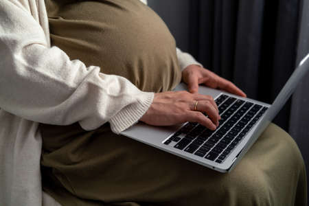 Attractive pregnant young lady working remotely online from home. Close up woman hands on laptop with big belly advanced pregnancy.の写真素材