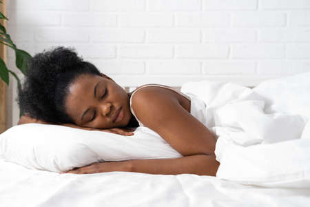 African American woman with hands under cheek sleeping in bed with white beddingの写真素材