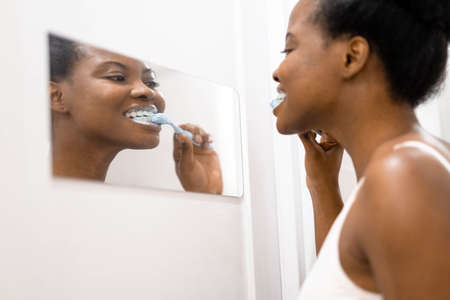 Portrait happy black African American girl brushing teeth in the bathroom. Morning routine with toothbrushes woman looking in mirrorの写真素材