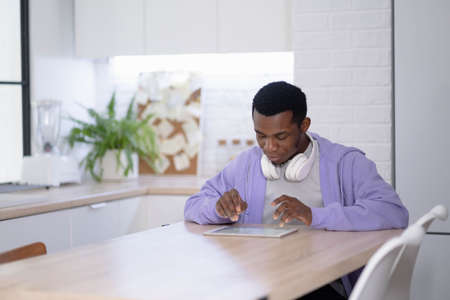 Black man using digital tablet on kitchen at home, working office work remotely typing text, African...