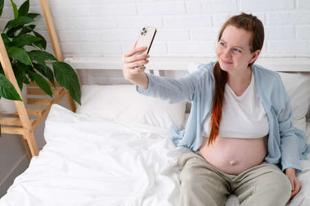Beautiful cheerful pregnant woman taking selfie photo using mobile phone lying on bed at home. big belly advanced pregnancyの写真素材