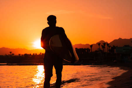 Silhouette man surfer in sunset light running down beach with surfboard near waters edge enjoying summer time practice hobby during getaway vacations, guy recreating at tropical island with wavesの写真素材