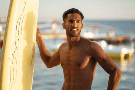 Portrait of young professional black surfer man, holds his surfboard in arm stands on ocean beach, sunset sun behind shoulder, sport hobby, relaxed lifestyleの写真素材
