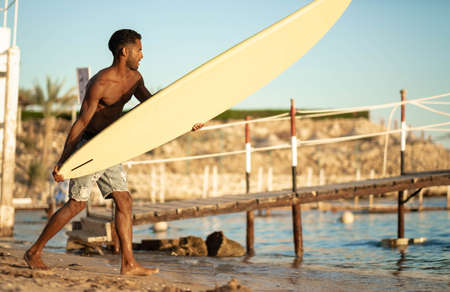 Portrait of young professional black surfer man, holds his surfboard in arm stands on ocean beach, sunset sun behind shoulder, sport hobby, relaxed lifestyleの写真素材