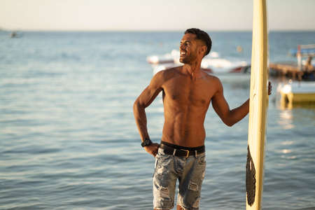 Portrait of young professional black surfer man, holds his surfboard in arm stands on ocean beach, sunset sun behind shoulder, sport hobby, relaxed lifestyleの写真素材