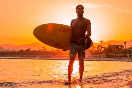 Silhouette man surfer in sunset light walking down beach with surfboard near waters edge enjoying summer time practice hobby during getaway vacations, guy recreating at tropical island with wavesの写真素材