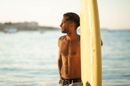 Portrait of young professional black surfer man, holds his surfboard in arm stands on ocean beach,...