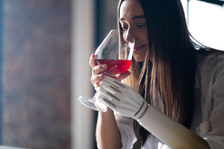 Calm thoughtful girl holds glass of wine in bionic arm at home, woman with artificial prosthetic limbの写真素材