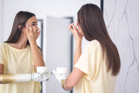 woman with prosthetic arm applying cream near mirror at home, girl with disability using artificial prosthetic limb to hold creamの写真素材