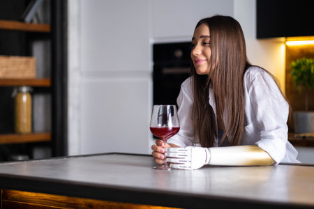 Happy girl with prosthetic arm holds glass of wine in artificial prosthetic limb hand at homeの写真素材