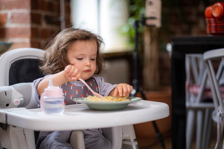 portrait caucasian baby girl about 2 years old in bib eating pasta from plate sitting high chair, self feeding for kids, baby-led weaning idea, healthy nutrition of solid food for infantの写真素材