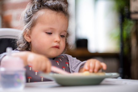 portrait caucasian baby girl about 2 years old in bib eating pasta from plate sitting high chair, self feeding for kids, baby-led weaning idea, healthy nutrition of solid food for infantの写真素材
