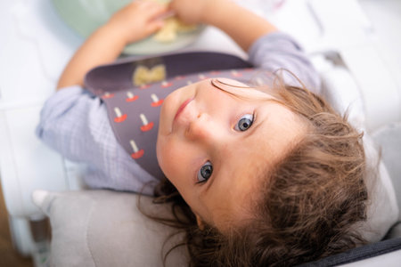 Top view portrait caucasian baby girl about 2 years old in bib eating pasta from plate sitting high chair, self feeding for kids, baby-led weaning idea, healthy nutrition of solid food for infantの写真素材