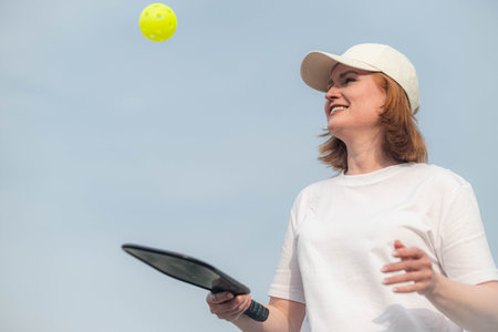 middle aged woman playing pickleball game over blue sky, pickleball yellow ball with paddle, outdoor sport leisure activityの写真素材