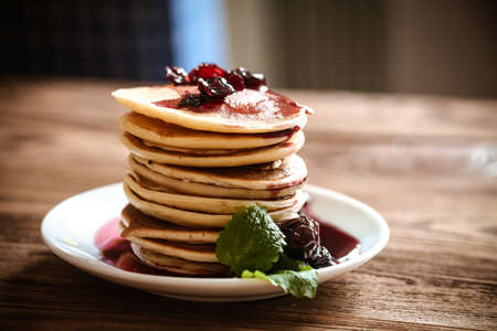 stack of pancakes on a white plate with cherry jam, mint leaves, brown wooden background of boards, rustic,の写真素材