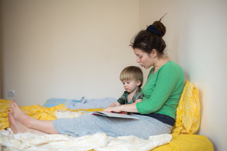Young mother and her son toddler reading book in a bedの写真素材