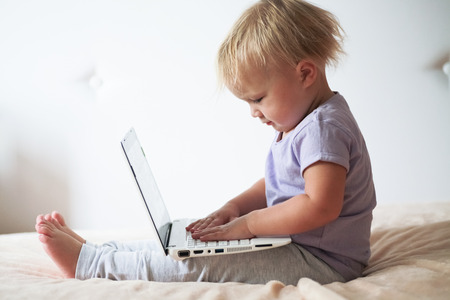 Adorable blond toddler  printing and  playing with laptop  pc at home, indoors. Concept children and gadgets.の写真素材