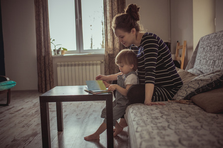 young pregnant mother with dreadlocks, and her son play and reading a book, lifestyle, real interior toningの写真素材
