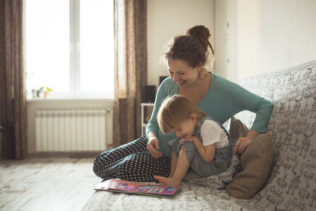 young pregnant mother with dreadlocks, and her son play and reading a book on sofa, lifestyle, real interior toningの写真素材