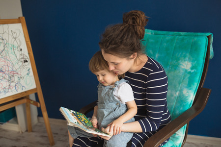 young pregnant mother with dreadlocks, and her son play and reading a book on sofa, lifestyle, real interior toningの写真素材