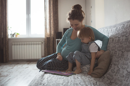 young pregnant mother with dreadlocks, and her son play and reading a book on sofa, lifestyle, real interior toningの写真素材