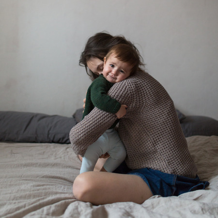 Shaggy young mother and baby in a knit sweater hugging and  playing on the bed, the concept home comfort, lifestyle, selective focus, toning, pastel colors, square formatの写真素材