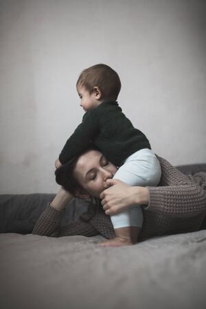Shaggy young mother and baby in a knit sweater hugging and  playing on the bed, the concept home comfort, lifestyle, selective focus, toning, pastel colorsの写真素材