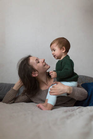 Shaggy young mother and baby in a knit sweater hugging and  playing on the bed, the concept home comfort, lifestyle, selective focus, toning, pastel colorsの写真素材