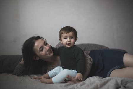 Shaggy young mother playing with baby on the bed, the concept of sweaters, comfort, lifestyle, selective focus, toningの写真素材