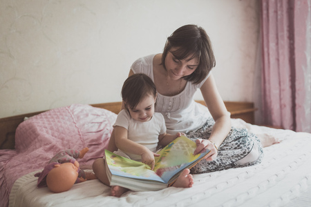 young dark haired mother and daughter Toddler reading a book sitting on the bed, lifestyle, pastel colors, selective focusの写真素材