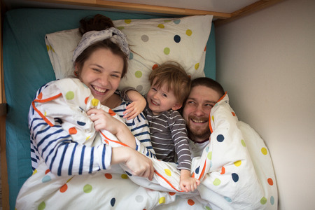 hugging a fun family with son playing in the children's bed, hiding under a blanket, casual, lifestyle, real interior, soft focus and light background, toningの写真素材