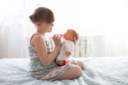 Kid girl playing with a doll, feeding a doll from a bottle, taking care of a doll, concept maternity, life style and childhoodの写真素材