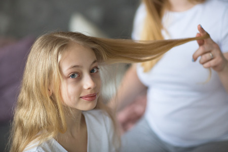 beautiful mother cares about a teenage daughter. Mom curls straightens hair to her daughter, Mom and daughter with long blond hair, concept of tenderness and care in real roomの写真素材