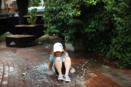 Happy funny baby toddler in a dress jumping barefoot in a puddle on the pavement, happy free childhood and summer walksの写真素材