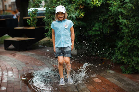Happy funny baby toddler in a dress jumping barefoot in a puddle on the pavement, happy free childhood and summer walksの写真素材