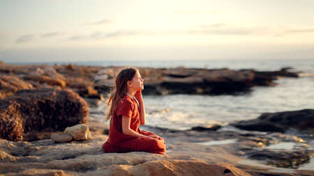 A European teenage girl in a dress sits on stones on the seashore, peace and quiet, a lonely child looks at the seaの写真素材